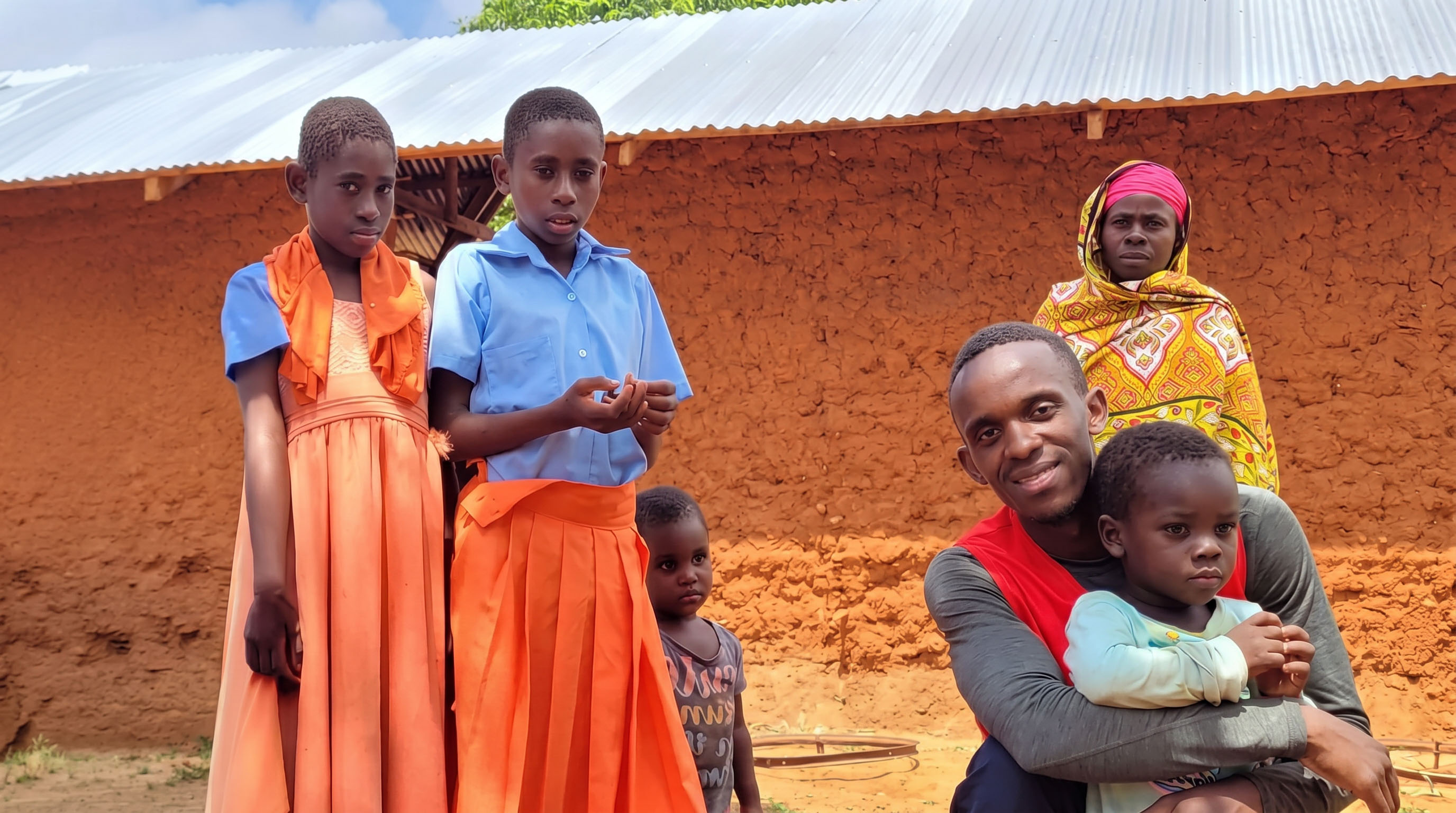 Ein lächelnder Helfer von Intena Humanity hält ein Kind im Arm, umgeben von Kindern und einer Familie vor ihrem Zuhause in Ostafrika
